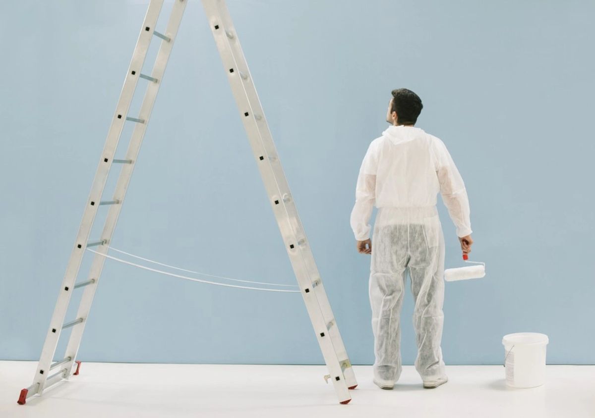 A painter in whites with a ladder and roller beside a freshly painted blue wall